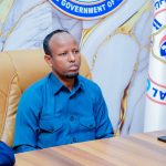 Wasiirka Caafimaadka oo xilka ka qaaday Agaasimihii Isbitaalka Hooyada & Dhallaanka Banaadir Nin fadhiyo kursi maamul oo ku labisan shaati buluug ah, hortiisna yaalla calanka iyo astaanta Wasaaradda Caafimaadka Soomaaliya.