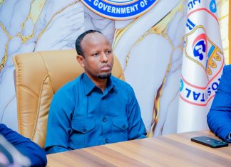 Wasiirka Caafimaadka oo xilka ka qaaday Agaasimihii Isbitaalka Hooyada & Dhallaanka Banaadir Nin fadhiyo kursi maamul oo ku labisan shaati buluug ah, hortiisna yaalla calanka iyo astaanta Wasaaradda Caafimaadka Soomaaliya.