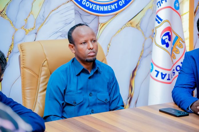 Wasiirka Caafimaadka oo xilka ka qaaday Agaasimihii Isbitaalka Hooyada & Dhallaanka Banaadir Nin fadhiyo kursi maamul oo ku labisan shaati buluug ah, hortiisna yaalla calanka iyo astaanta Wasaaradda Caafimaadka Soomaaliya.